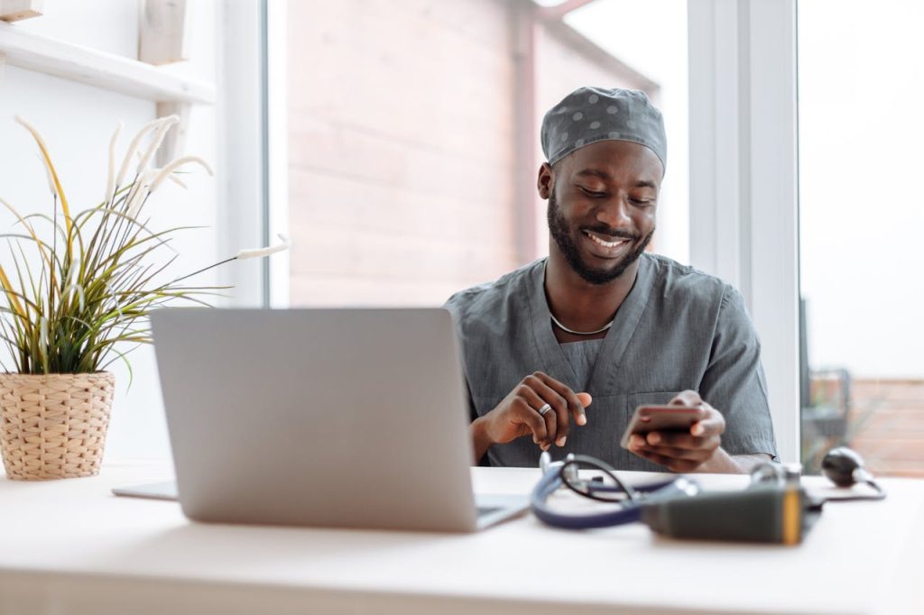 pexels photo 5327914 Smiling healthcare professional using phone in office with laptop and medical tools.