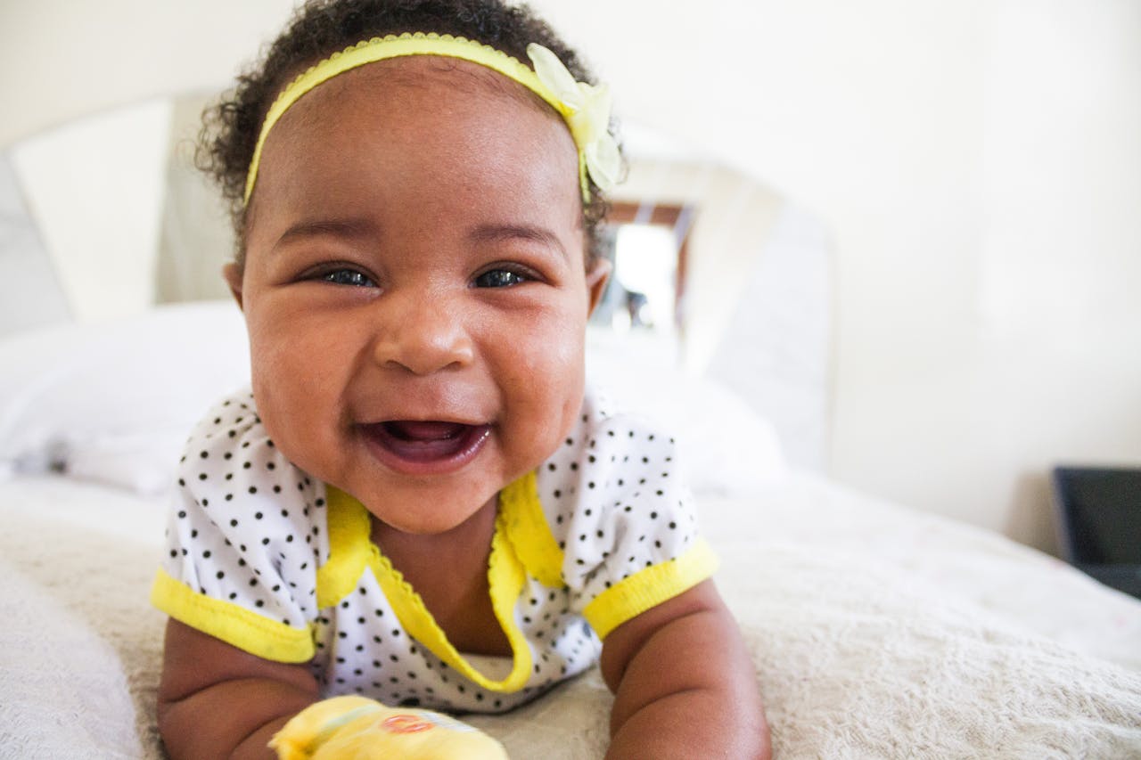 why-choose-us Cheerful baby girl in a polka dot outfit with a big smile on a bed indoors.